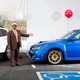 A car dealer in a brown suit holding his arm outstretched toward a blue Subaru with a red balloon tied to it