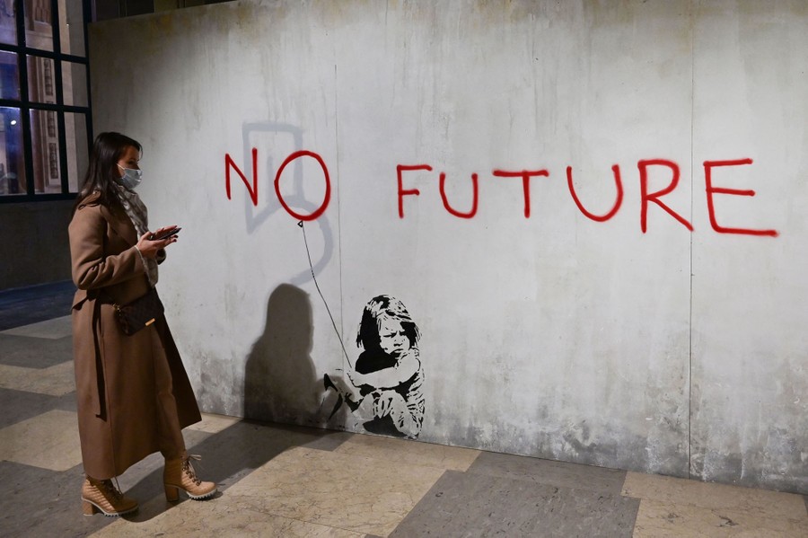 A woman stands beside a mostly blank wall that features a stencil of a sitting girl and has the words "No Future" painted on it.