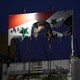 A tattered and torn billboard of the Syrian flag with Bashar al-Assad’s face on it lit up atop a building at night