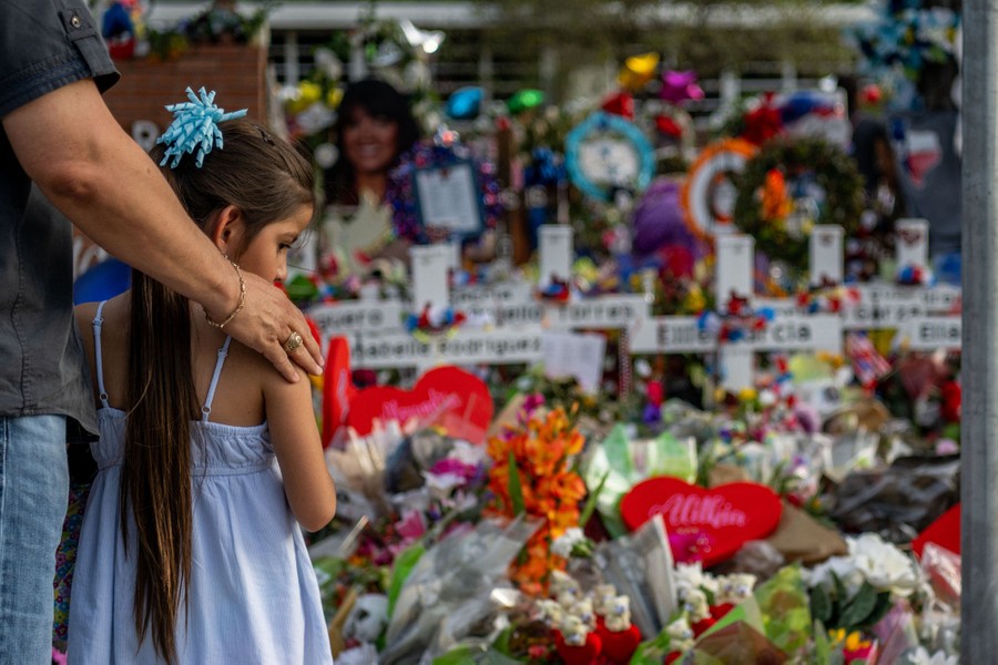 A father and his young daughter stand together in front of a memorial made up of temporary crosses and many bouquets of flowers.