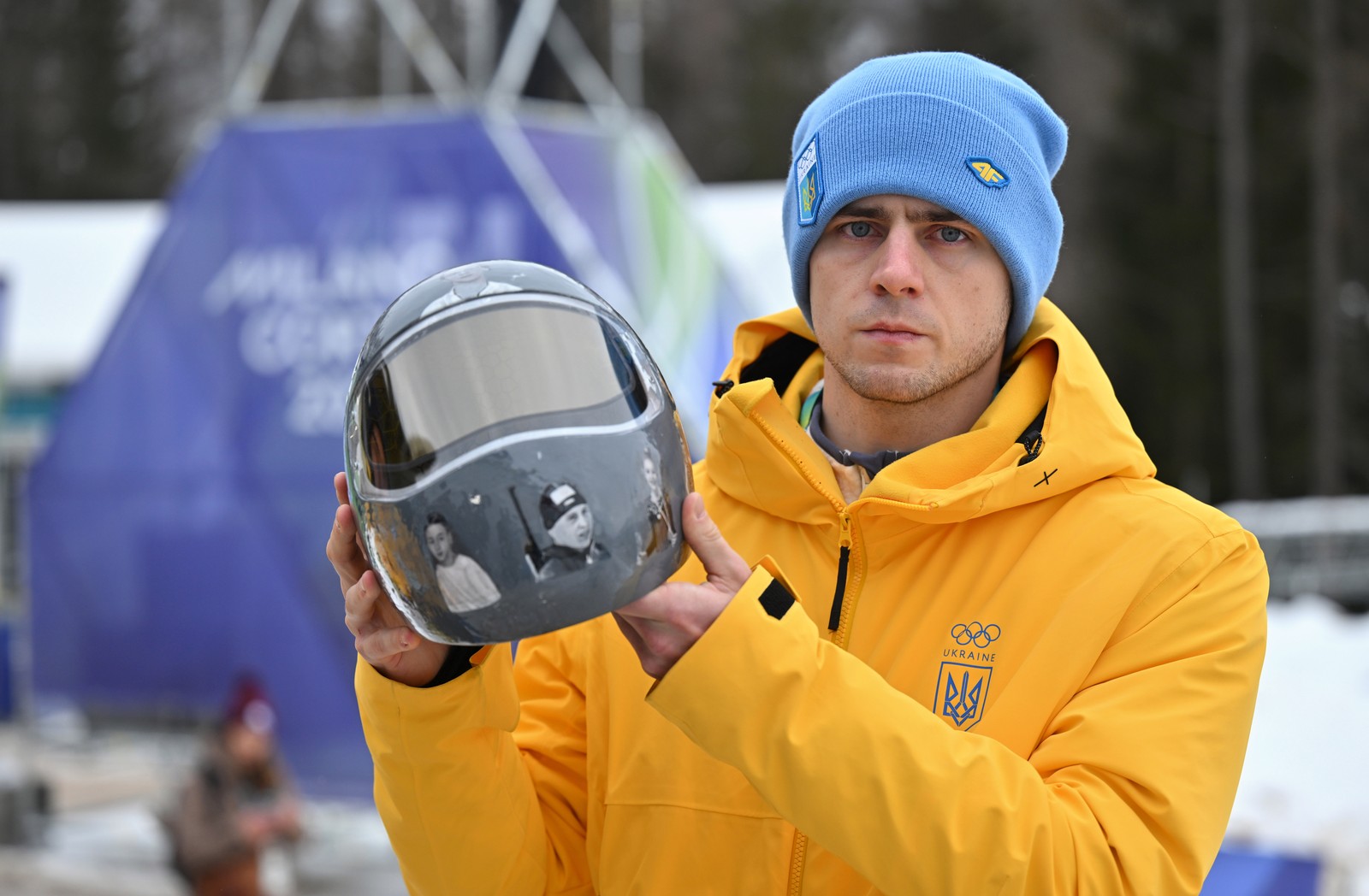 A grim-looking athlete wearing a blue beanie and yellow jacket holds up a gray racing helmet adorned with many images of other athletes.