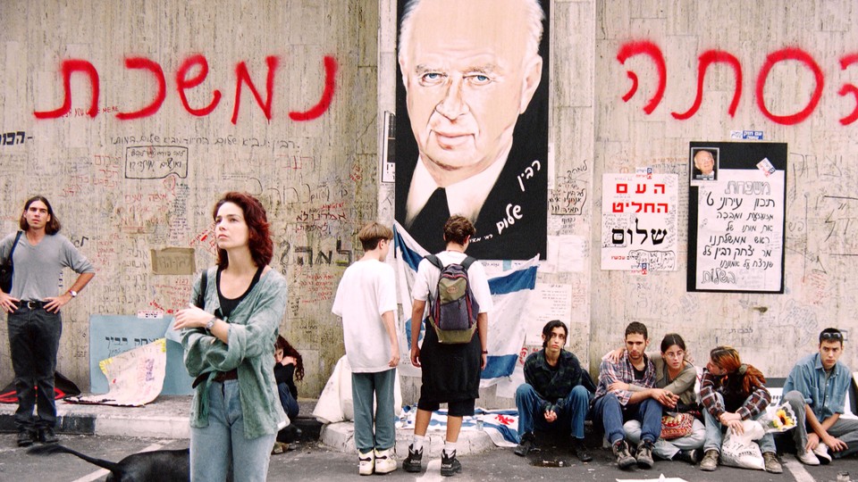 Photograph of a wall with an illustration of Yitzhak Rabin, Hebrew writing, and mourning protesters sitting in front of it