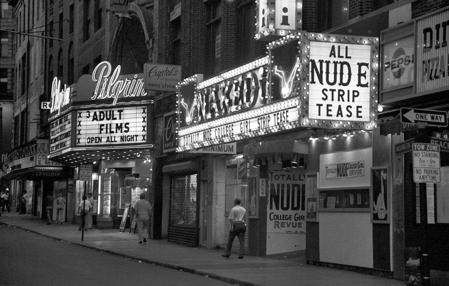Several people walk past a strip club and an adult-film theater on a city street.