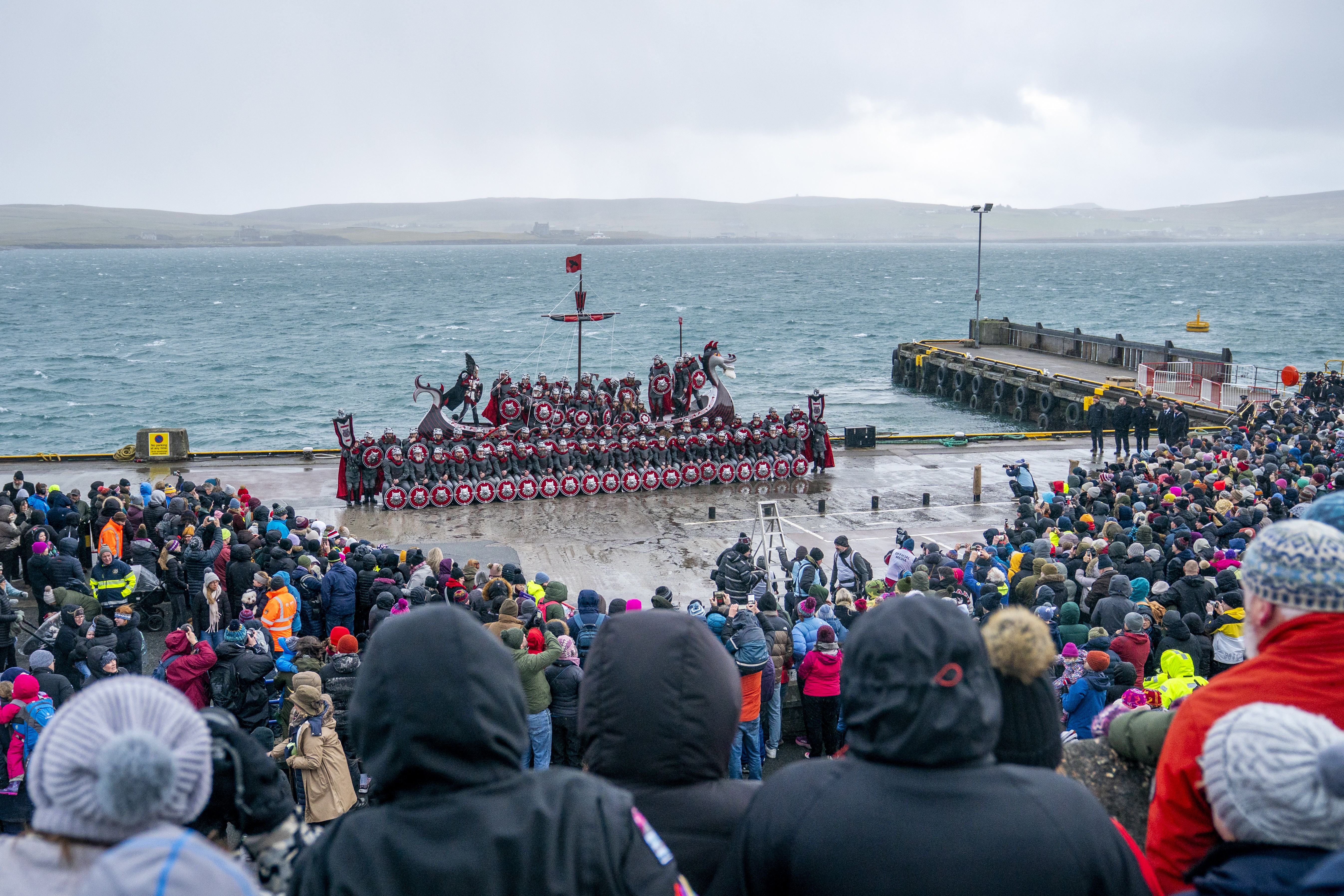 A crowd gathers around a group of people dressed as Vikings who pose beside a replica of a Viking ship beside a harbor.
