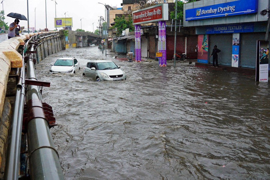People on an overpass look below them as two cars are pushed along a flooded street by the current.