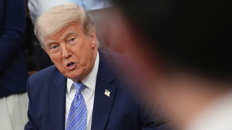 Photograph of a seated Donald Trump speaking. The image is taken from the behind the left shoulder of a blurred male figure.