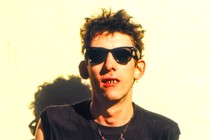 Shane MacGowan standing against a cream and green wall wearing a Pogues black shirt and sunglasses