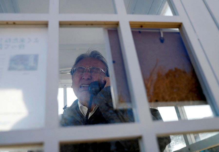 A man stands inside a white glass-and-wood phone booth, emotional, while speaking into a receiver.