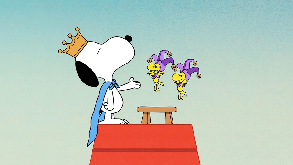 Atop his doghouse, Snoopy wears a crown and a cape as two small yellow birds in jester costumes hover.