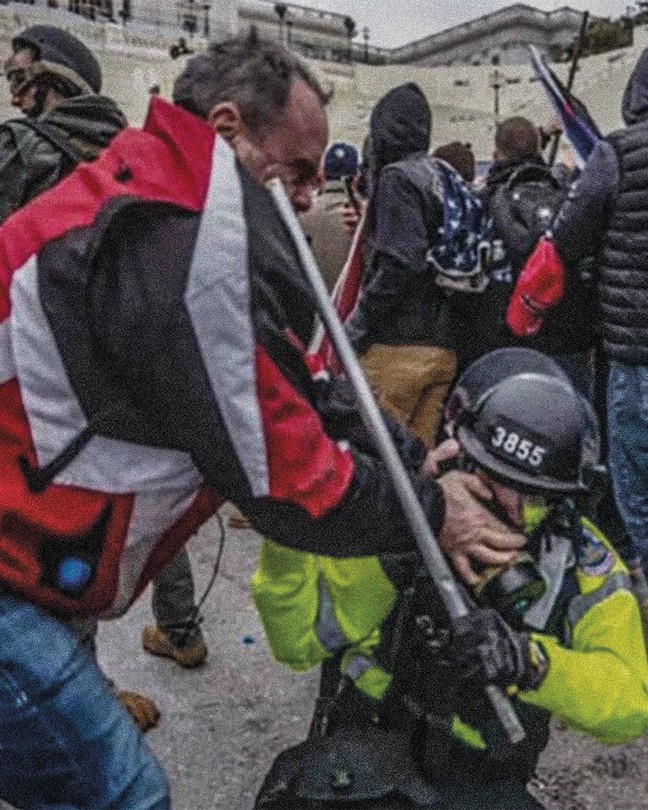 photo of man in red, gray, black jacket and jeans standing over and pushing his thumbs into the eyes of a kneeling police officer in helmet and gear