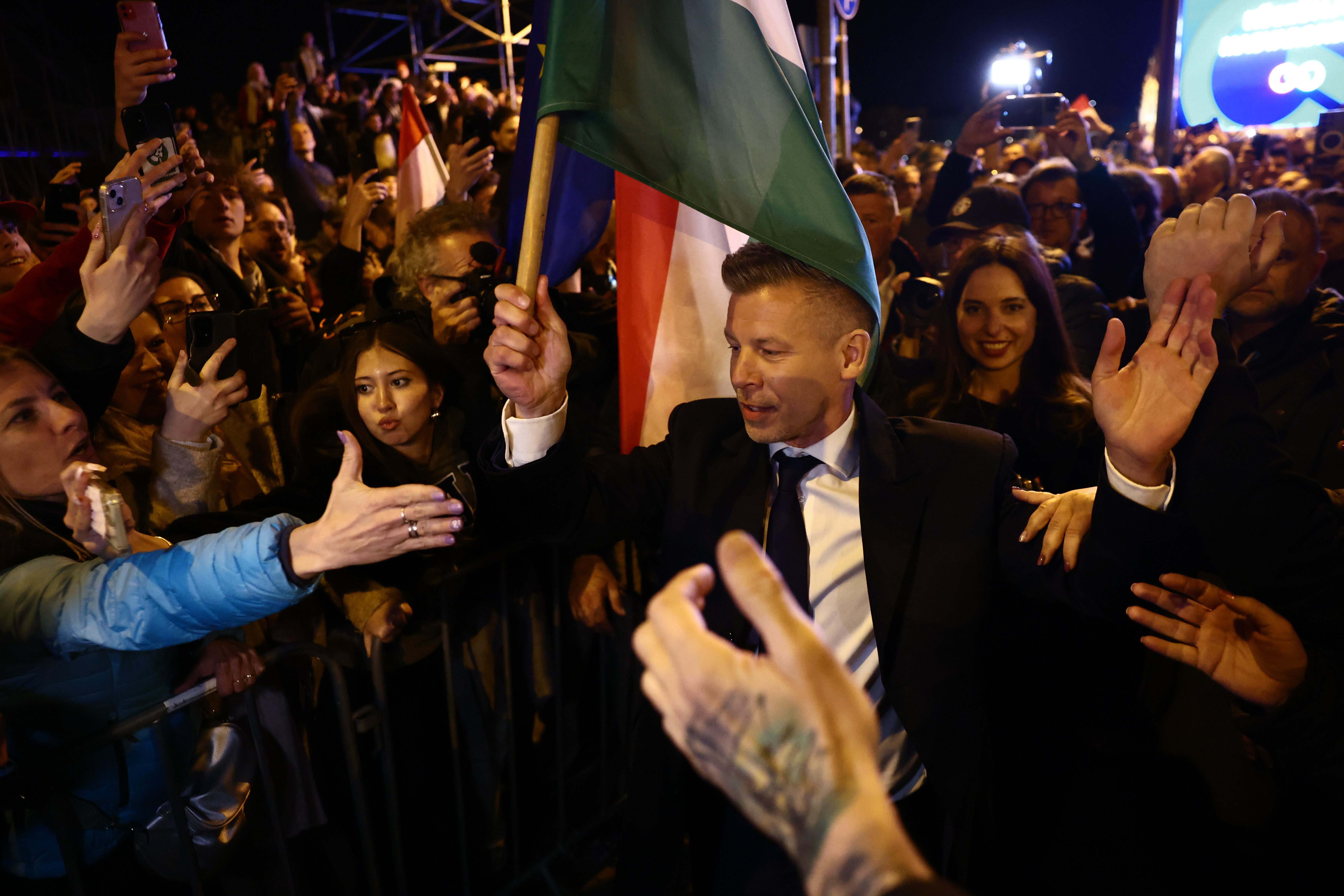 Supporters in a crowd cheer, take photos, and reach out as a politician waving a Hungarian flag walks past them.