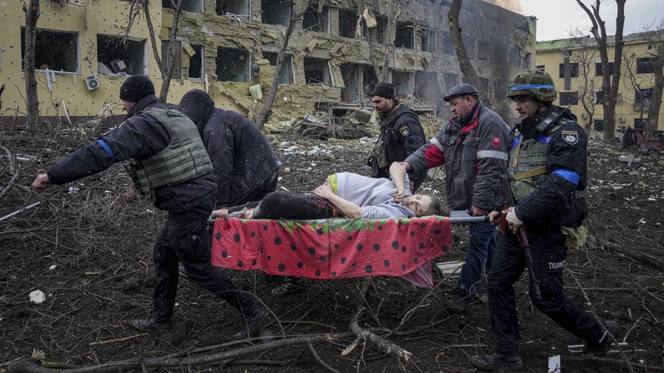 Five men carry a pregnant woman on a stretcher. They are surrounded by destroyed buildings and debris.