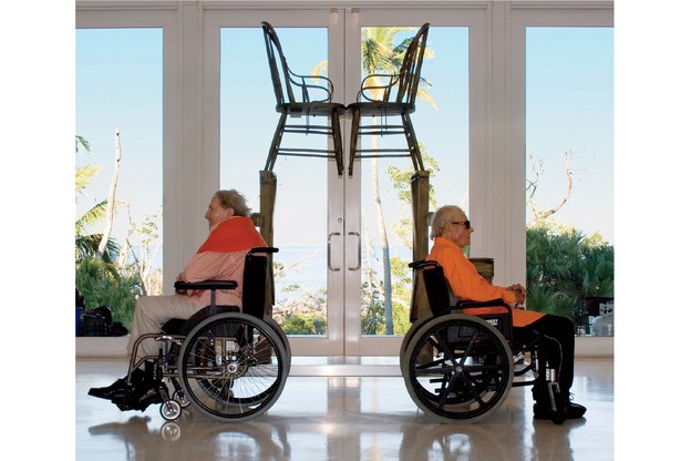 photo of two men in wheelchairs facing away from each other, each in front of the mirrored stair-step portions of Rauschenberg's 1981 sculpture, with the facing chairs visible at top and window overlooking palm trees and ocean behind