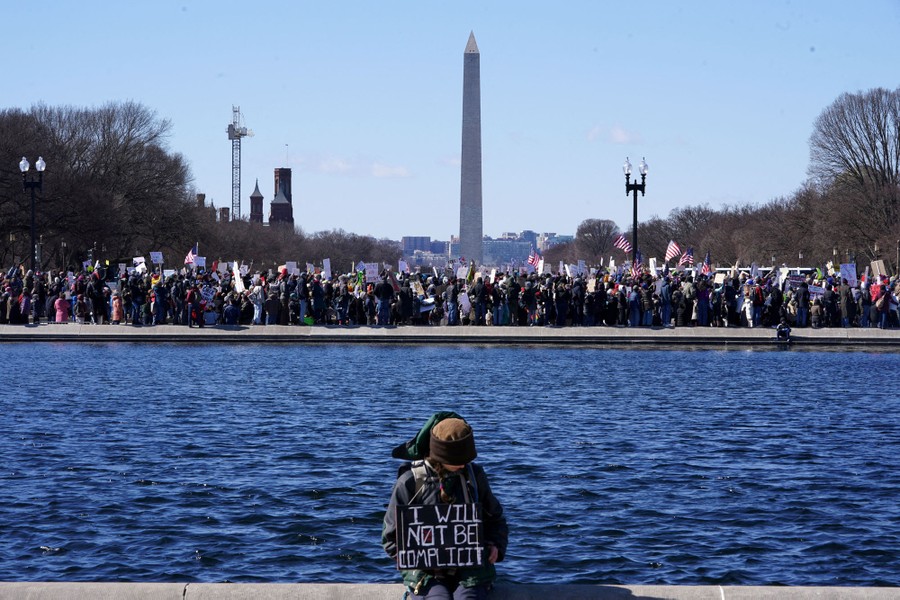 A single protester sits along a reflecting pool on the National Mall, holding a sign that reads 