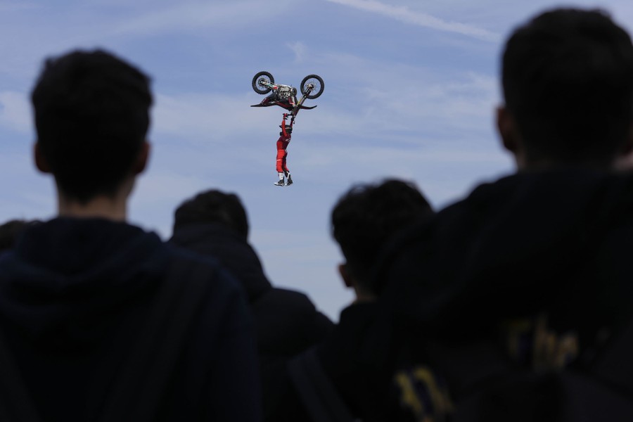 People watch a stunt performer who hangs from the handlebars of an upside-down motorcycle in midair.