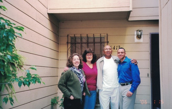Two older women, one older man, and a young man stand with their arms around one another in front of a house