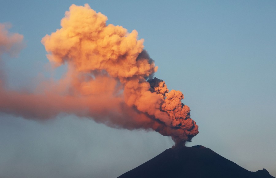 A cloud of ash rises from a volcano, catching the colors of the sun low in the sky.
