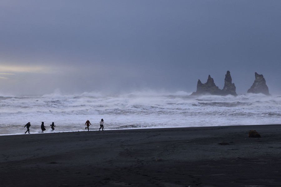 Five tourists run on a black-sand beach, with waves crashing beyond.