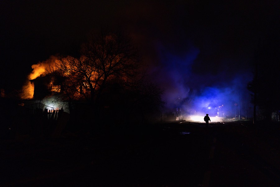 A firefighter in silhouette at night on the right side of the image, and a burning house on the left