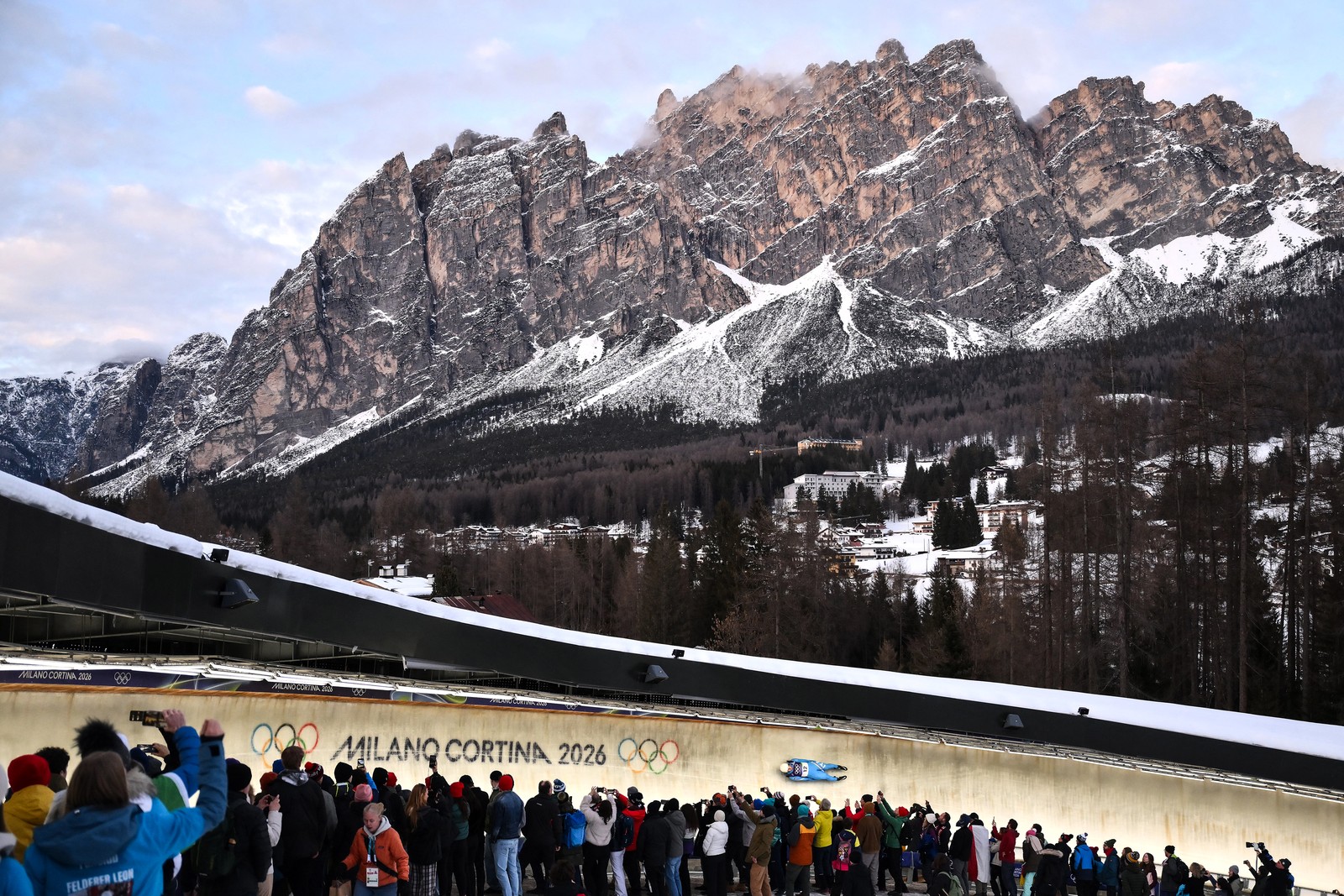 A luge rider races on a curving ice track in a resort town beneath a towering mountain.