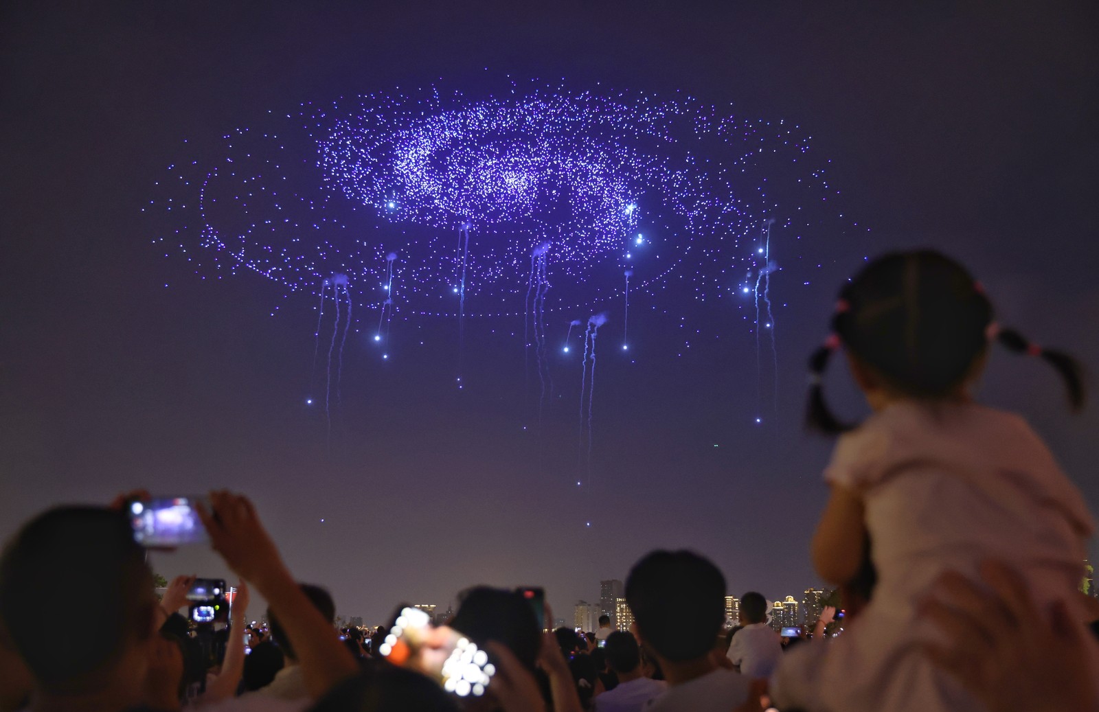 People look up toward a drone show which is creating the shape of a spiral galaxy in the sky above them.