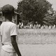 Photograph of an African American boy watching a group of people, some carrying American flags, marcing past to protest the admission of the "Little Rock Nine" to Central High School