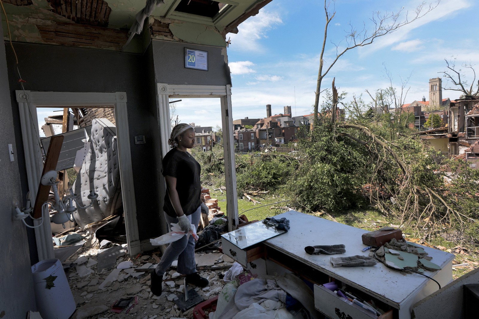 A person walks through a house that is missing an entire wall, with storm damage visible outside.