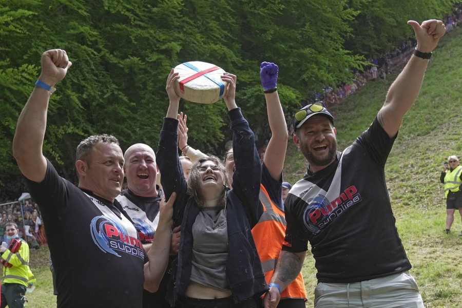 A woman holds a wheel of cheese above her head, surrounded by a group of cheering people.