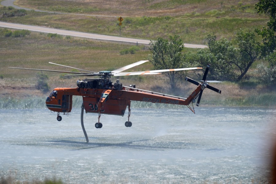 A helicopter hovers above a small lake, lowering a hose into the water to fill up for a drop on a nearby fire.