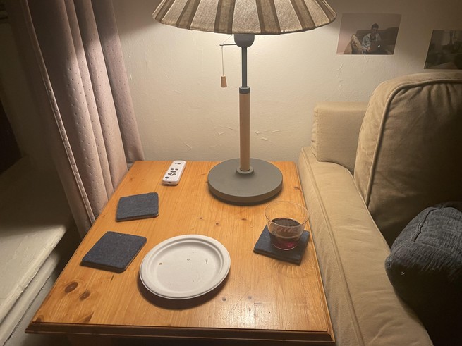 A side table with a glass of wine and a lamp, in front of a wall with snapshots taped to it.