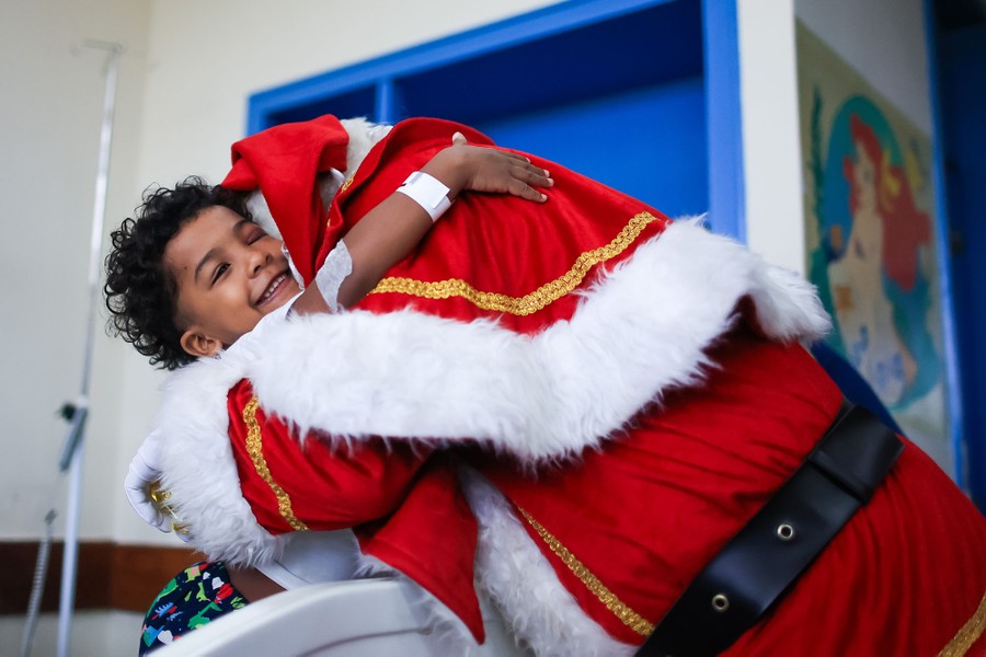 Dressed as Santa Claus, a person leans over to give a big hug to a young patient in a hospital bed who smiles and hugs back.