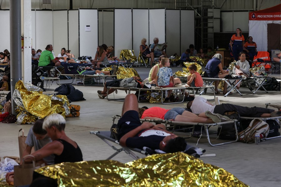 A couple dozen people sit and lie on cots inside a large warehouse-like space.