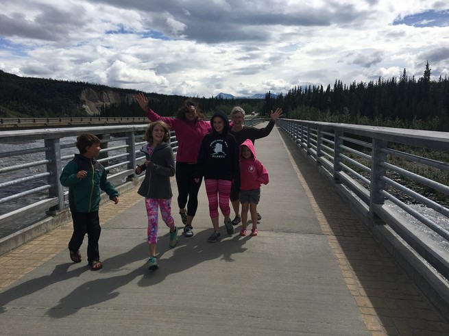 Two women and four children run across a bridge with a forest in the background, under a blue, cloudy sky