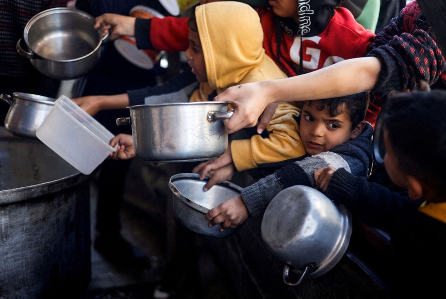 Children stand at the front of a crowd that pushes forward, holding out bowls and pots to receive food.