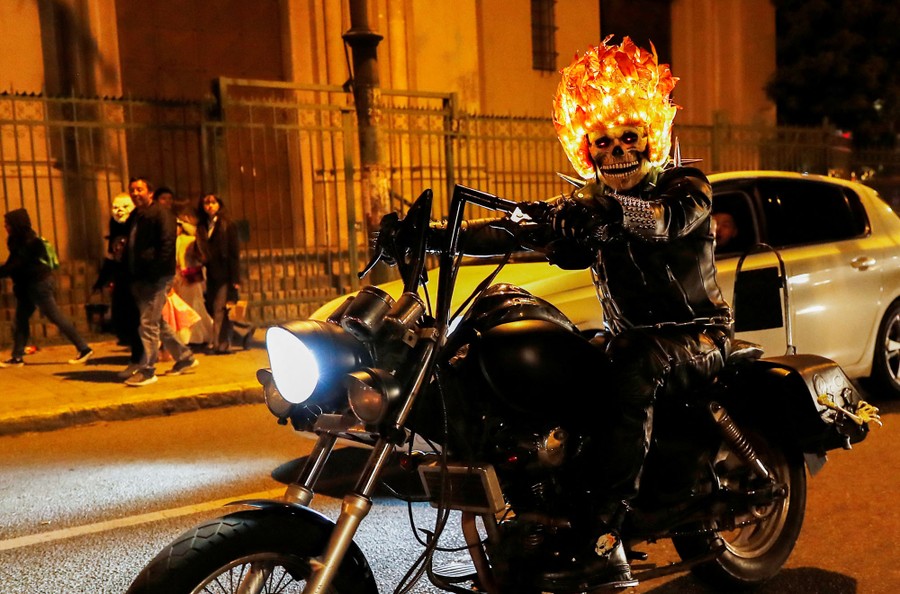 A person rides a motorcycle while wearing a skull mask and an illuminated helmet made to look like flames.