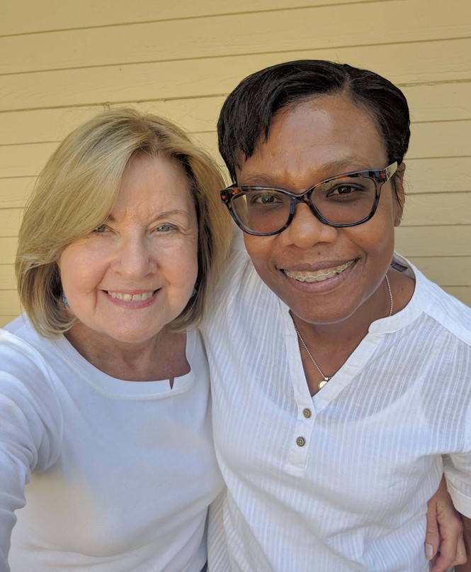 Two women embrace in a close-up selfie; the one on the left is an older white woman with a blond bob, the one on the left is a middle-aged Black woman with glasses, braces, and a pixie cut