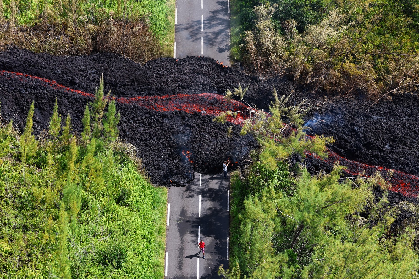 An aerial photograph shows a lava flow cutting across a paved road in a forested area.