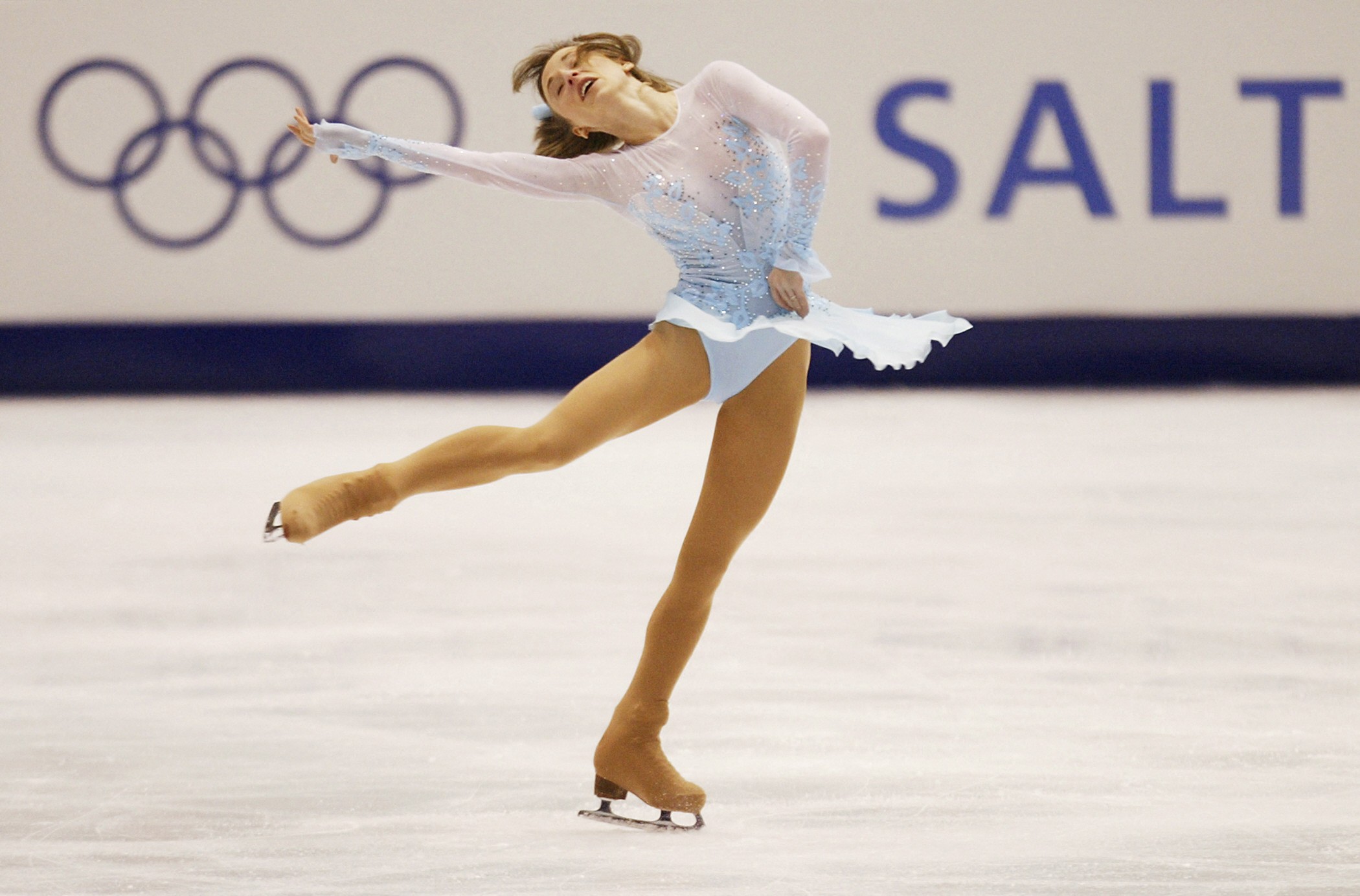 photo of woman in white short-skirted skating costume performing on ice rink with Olympic logo in background