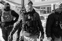Black-and-white photograph of four ICE agents wearing caps and face masks on a sidewalk in a suburban neighborhood