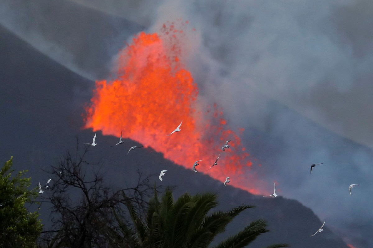 Pigeons fly in front of a fountain of erupting lava.