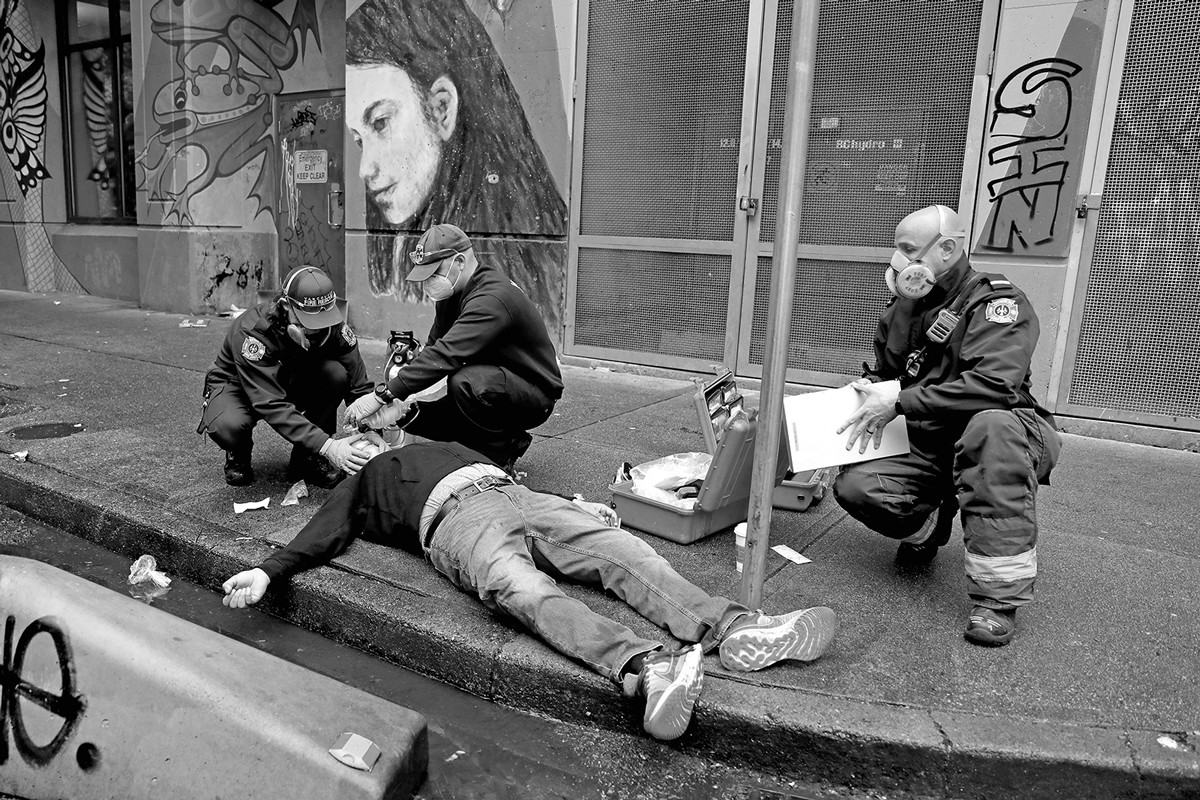 black-and-white photo of overdosing man lying on sidewalk curb with three people attending