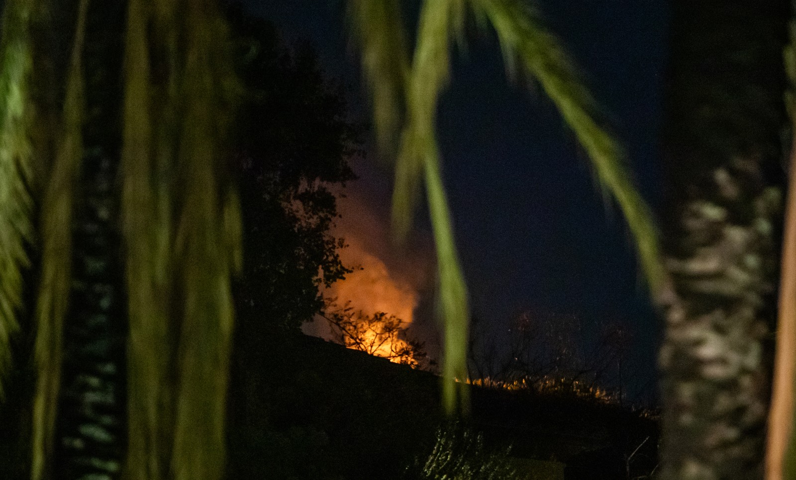 Light and smoke from a small fire, seen on a hillside at night.