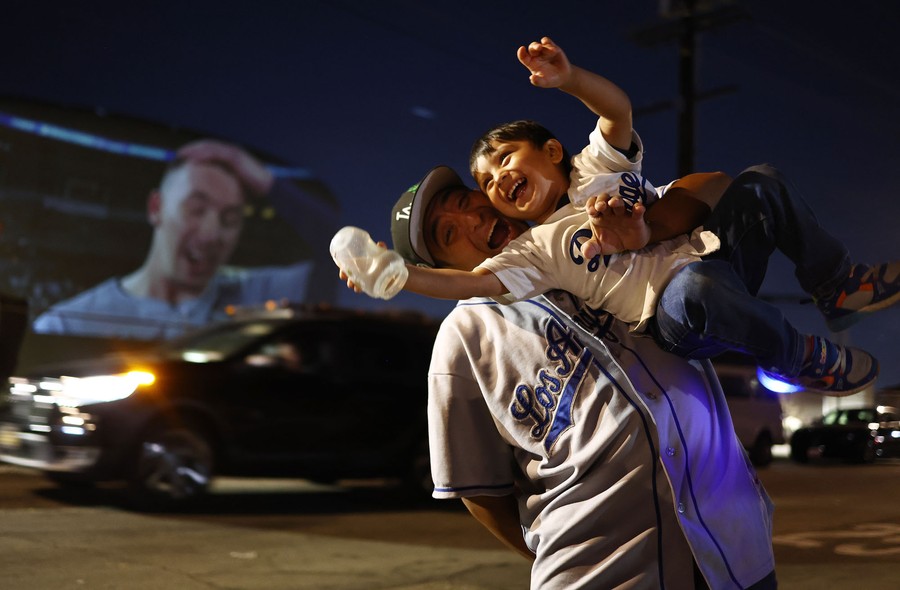 A man smiles and lifts his son up, celebrating while standing in front of a projected screen showing a baseball player.