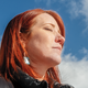 photo of red-haired woman with eyes closed wearing black jacket in sunlight with mountain and blue sky in background