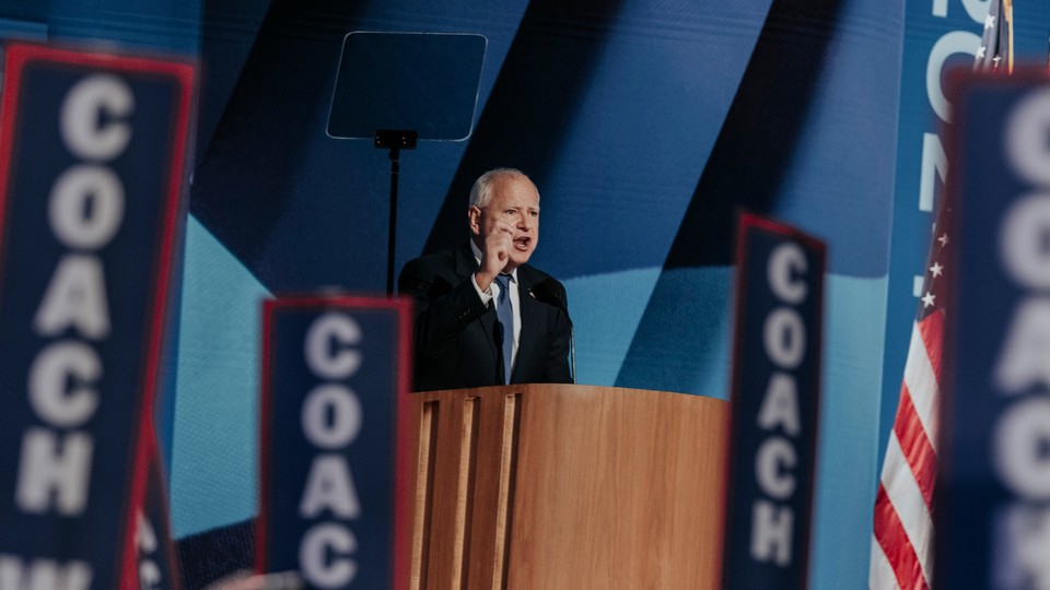 A man in a suit and blue tie with balding white hair speaks from a podium with signs that say coach around him.