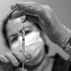 A black and white photo of a woman wearing a mask pulling the COVID-19 vaccine into a syringe with gloved hands