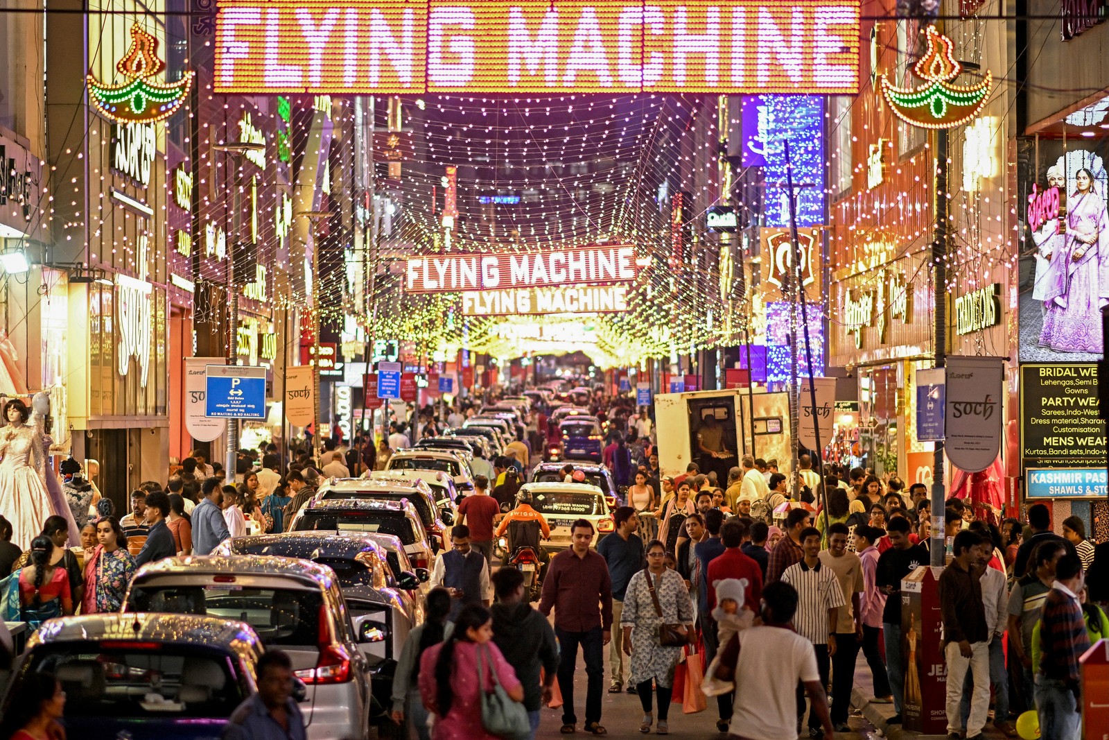 Crowds of people walk through a shopping district in a city that is lit with many hanging lights.