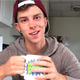 Person wearing backwards red baseball cap and gray sweatshirt holding a mug and looking at camera