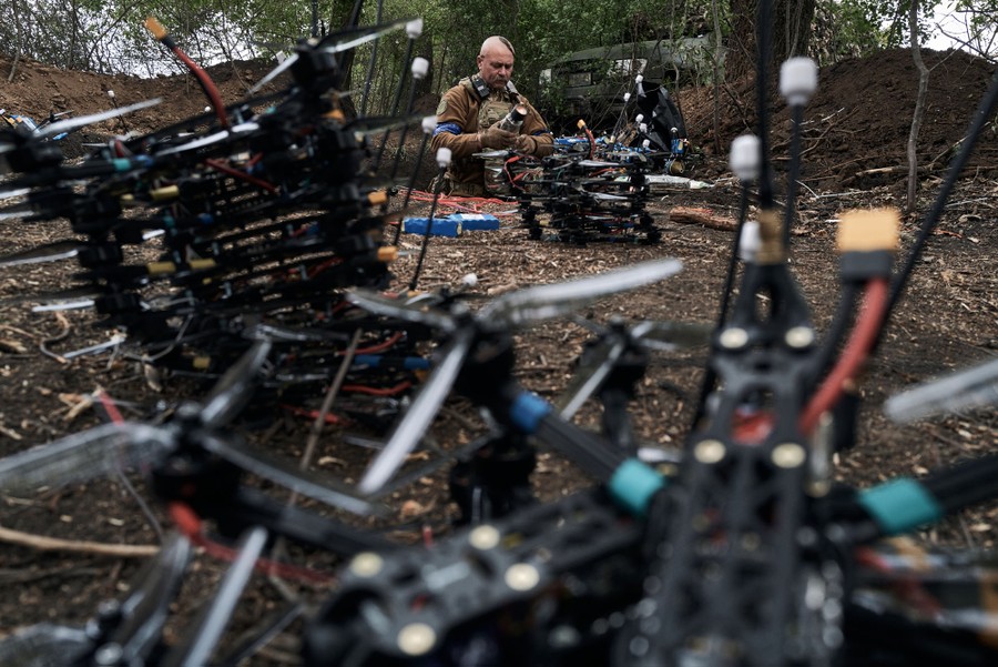 A soldier in a trench works on one of dozens of small four-rotor drones laid on the ground around him.