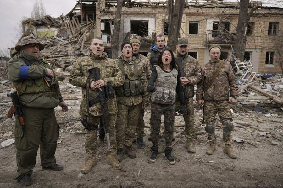Eight people wearing varied military uniforms stand together amid rubble, singing.